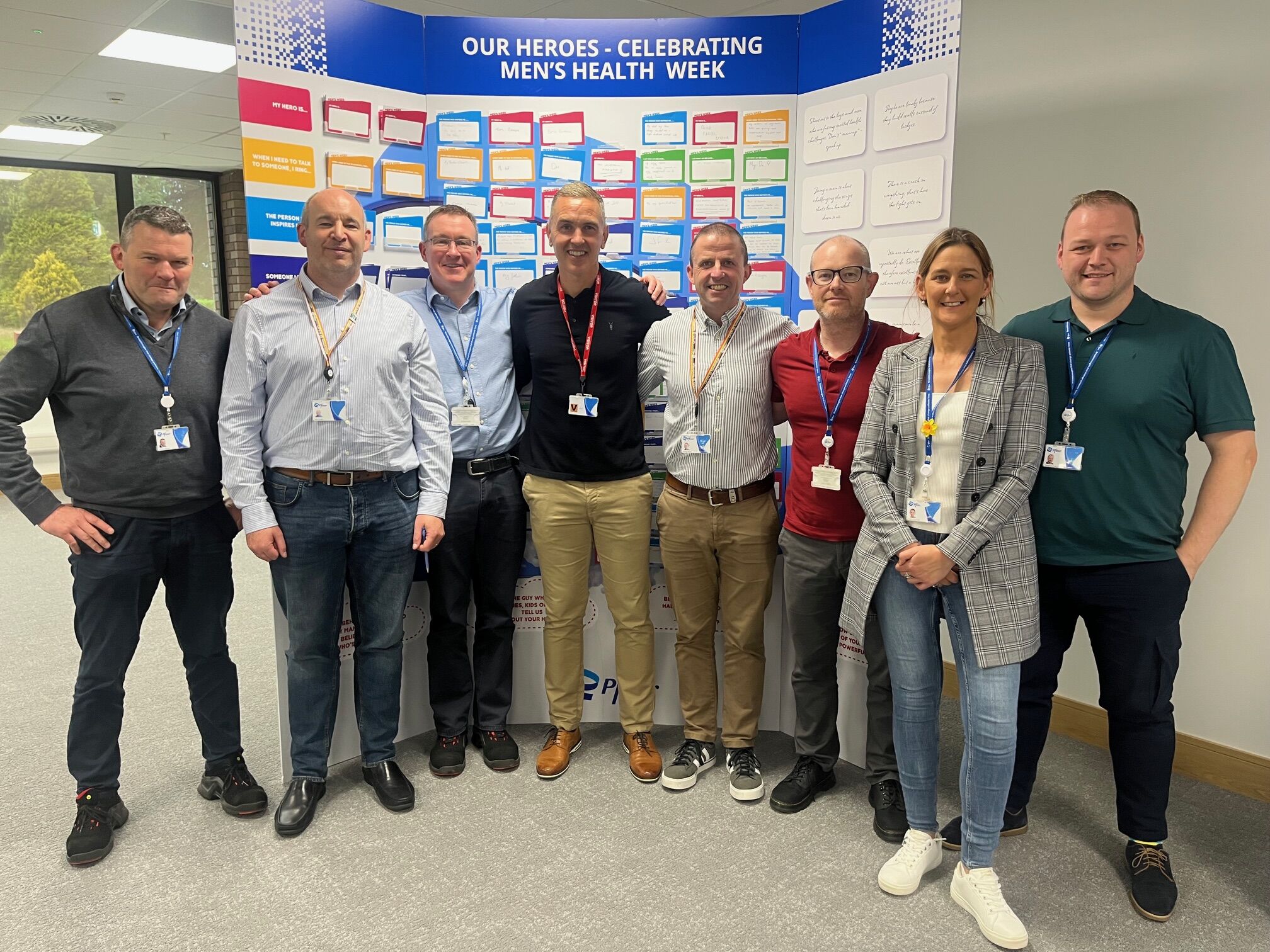 Colleagues posing together in front of a "Our Heroes - Celebrating Men's Health Week" display board